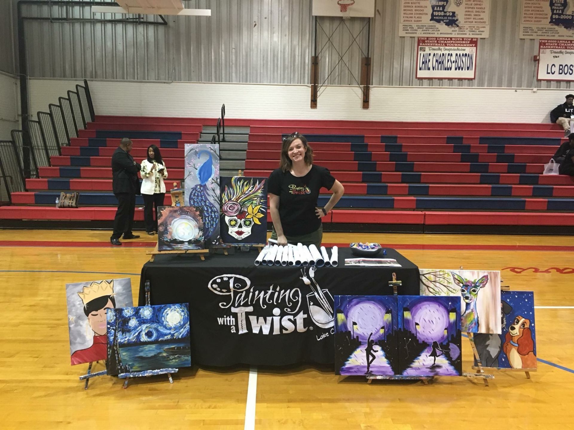 A woman is standing in front of a table with paintings on it in a gym.