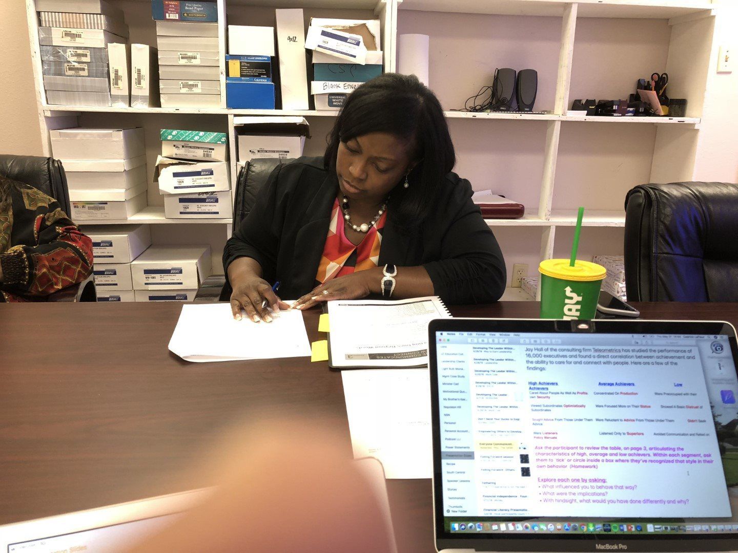 A woman is sitting at a desk with a laptop and a green cup that says nbv on it
