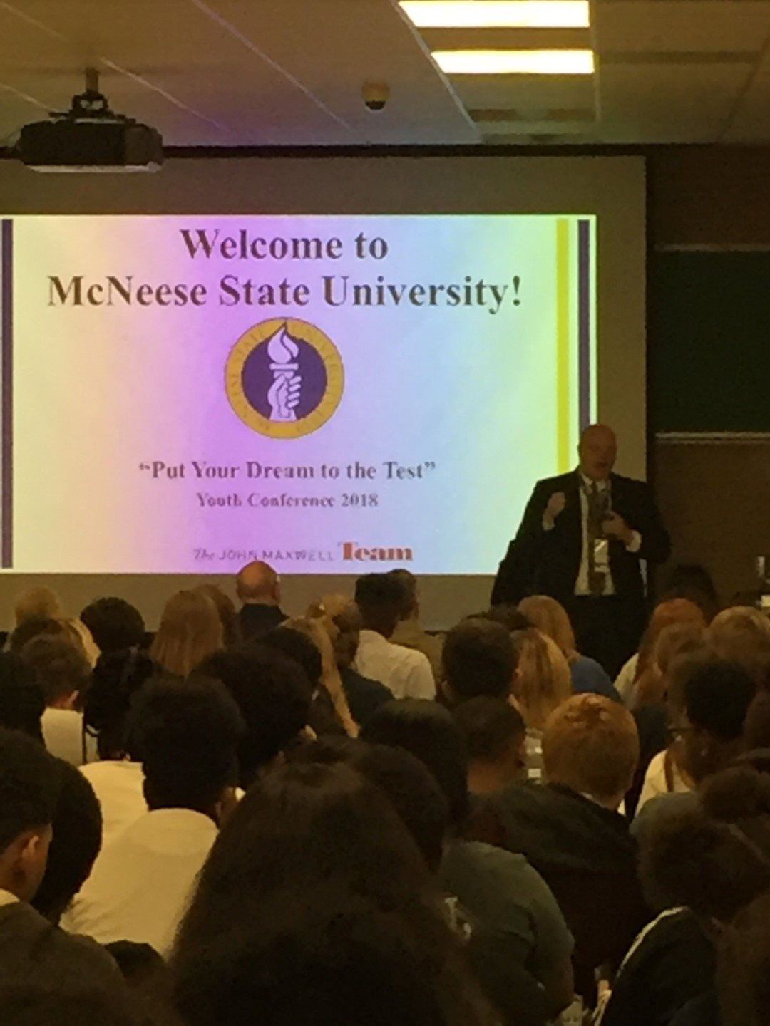 A man stands in front of a projector screen that says welcome to meneese state university