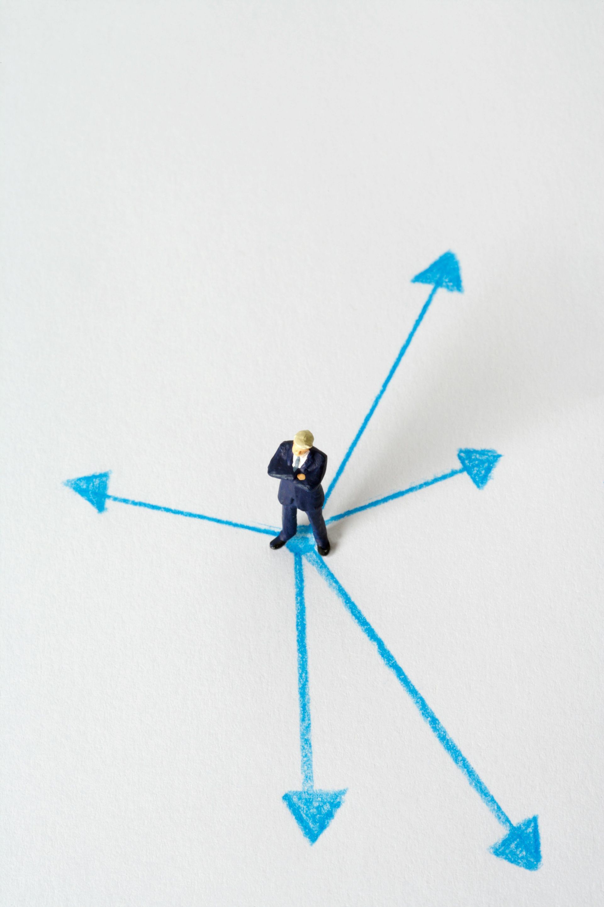 Miniature businessman standing at the intersection of five blue arrows on a white surface.