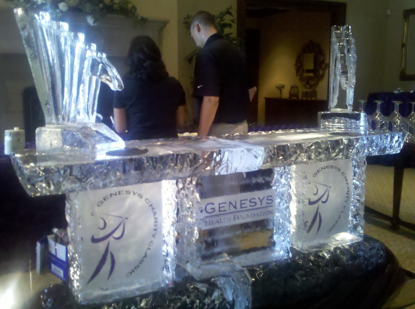 A man and a woman stand behind a bar made of ice that says genesys