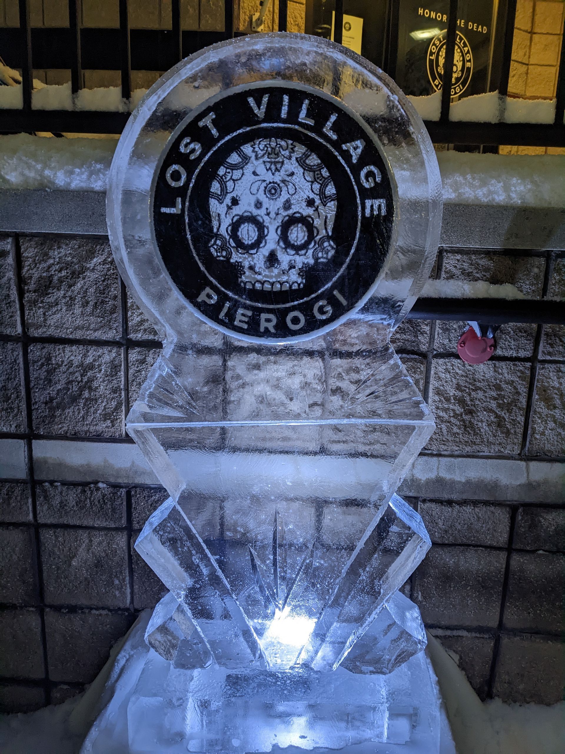 A lost village pierogi ice sculpture is sitting in front of a brick wall.