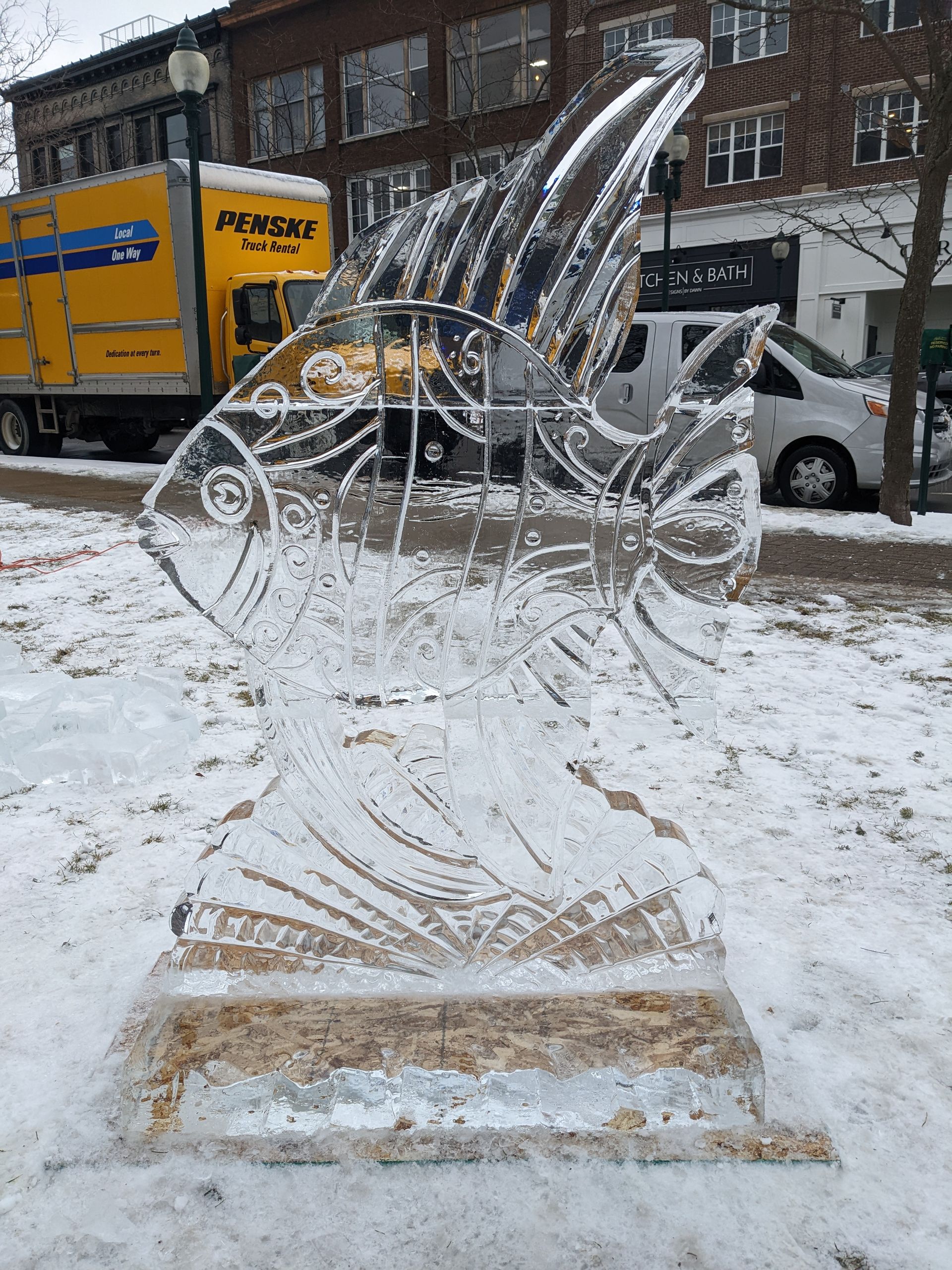 A large ice sculpture of a fish is sitting in the snow in front of a moving truck.