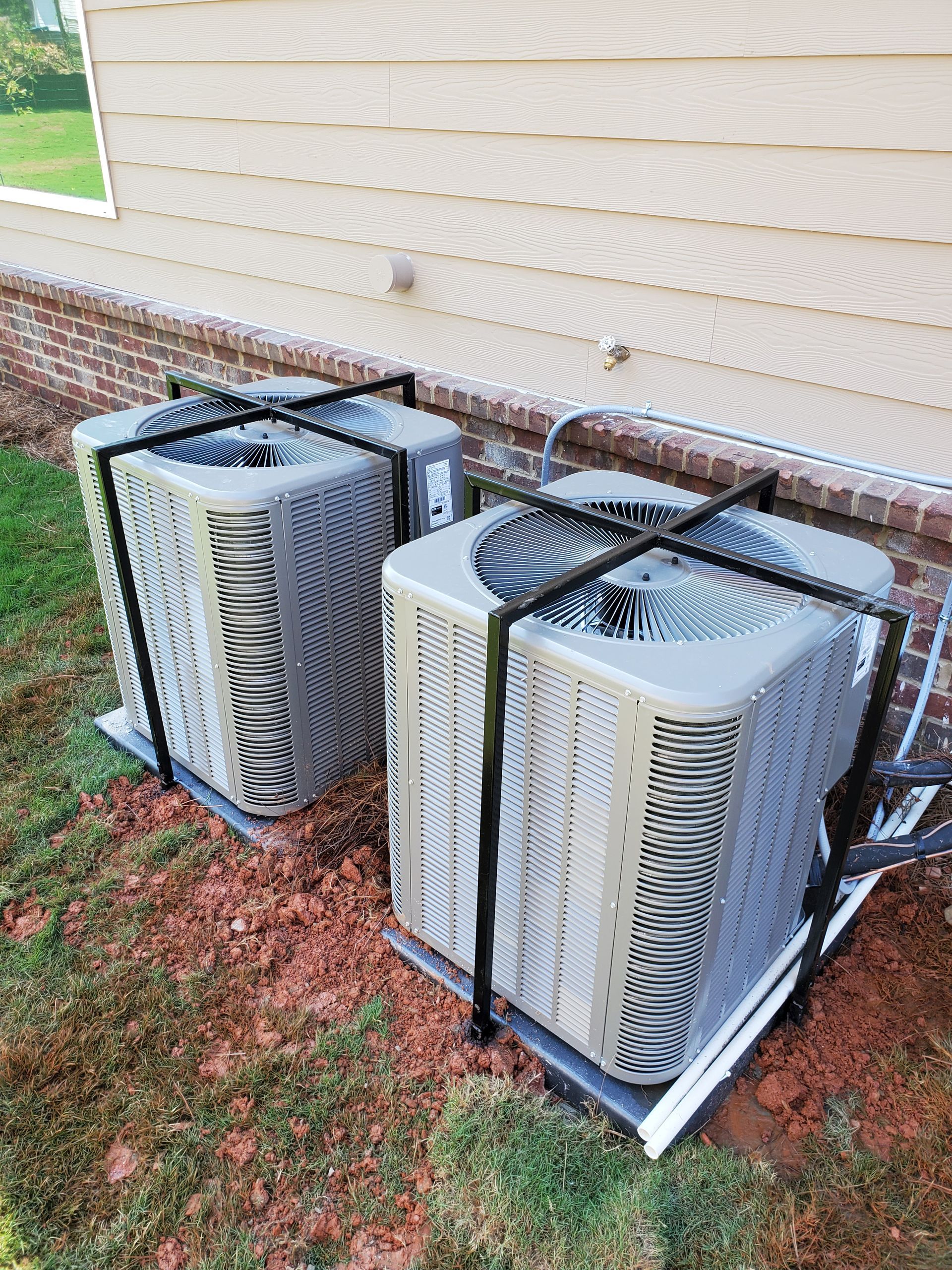 Two air conditioners are sitting next to each other on the side of a house.