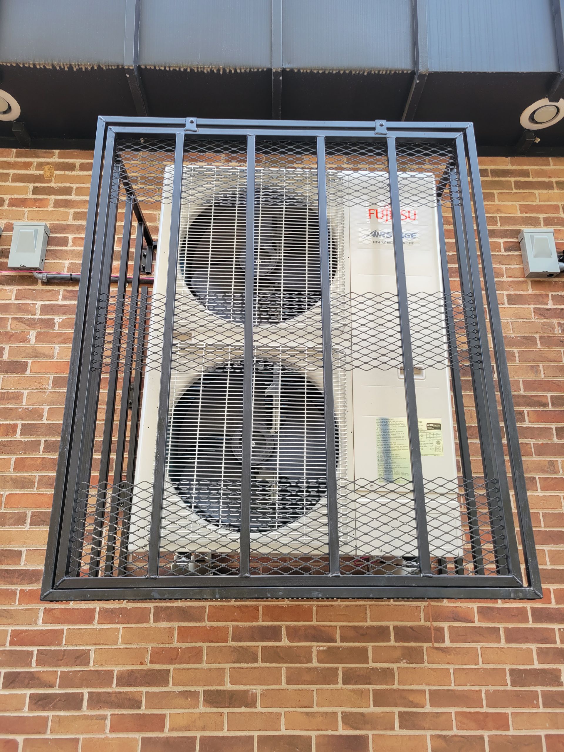 An air conditioner is behind a metal fence on a brick wall.