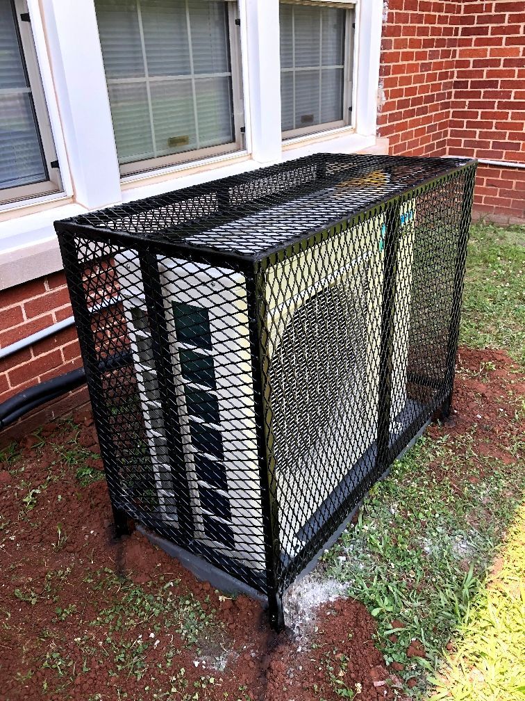 An air conditioner is sitting in a metal cage in front of a brick building.