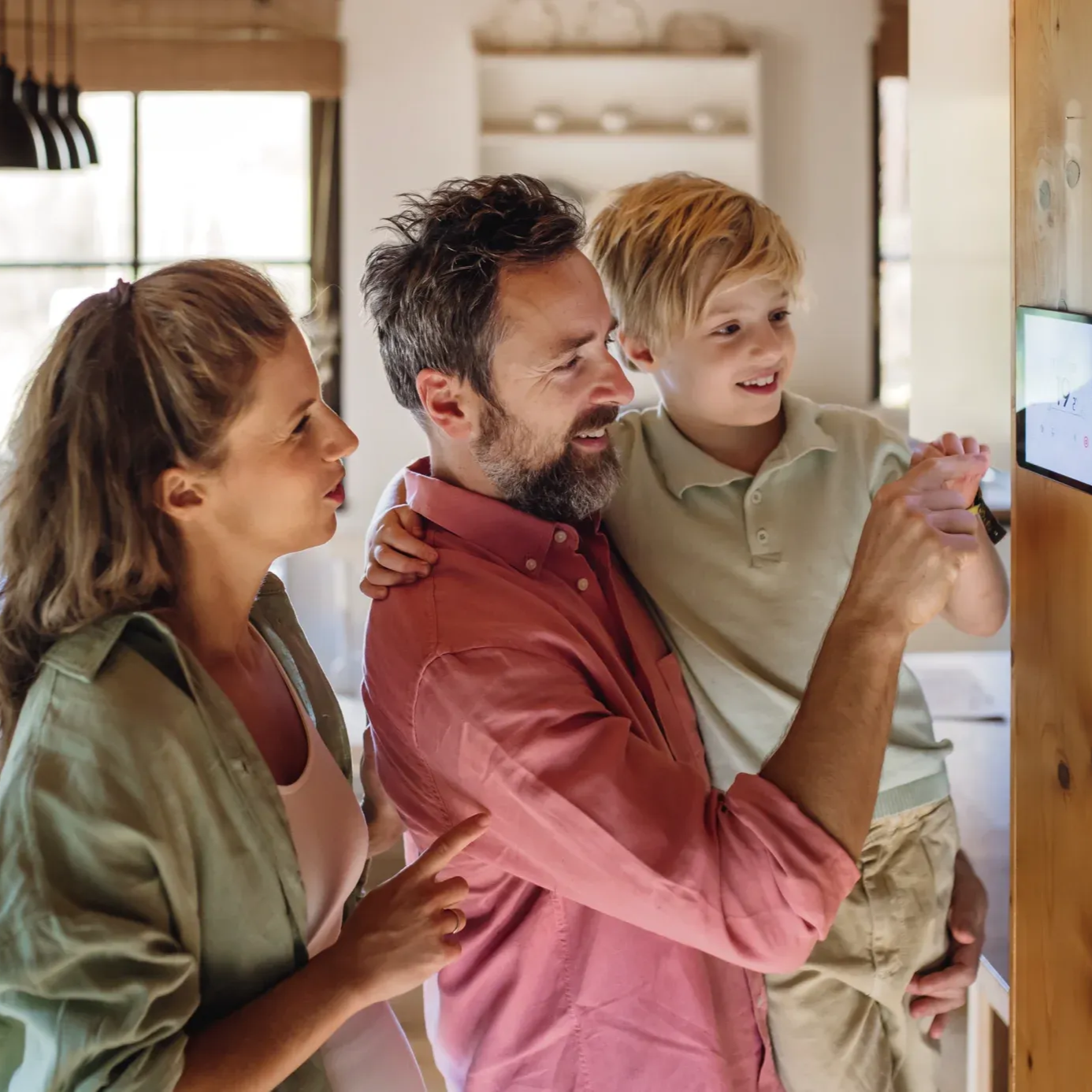 A family of three interacts with a smart home display mounted on a wooden wall in a brightly lit, modern kitchen.