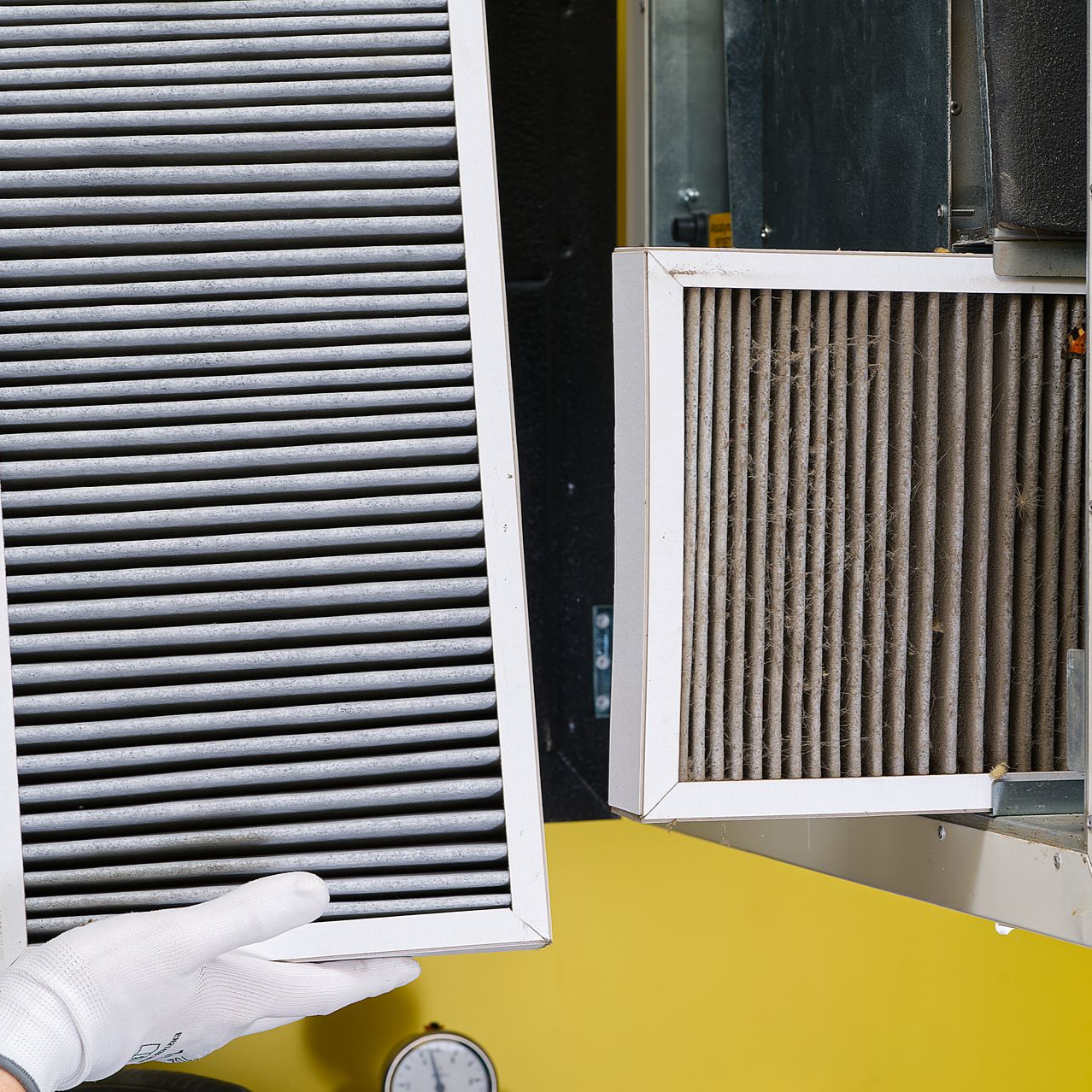 A gloved hand holding a new, dark gray air filter next to a dirty, used one installed in an HVAC system.