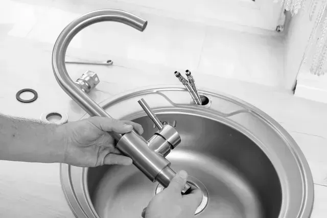 Hands holding a metal kitchen faucet above a stainless steel sink, preparing for installation.