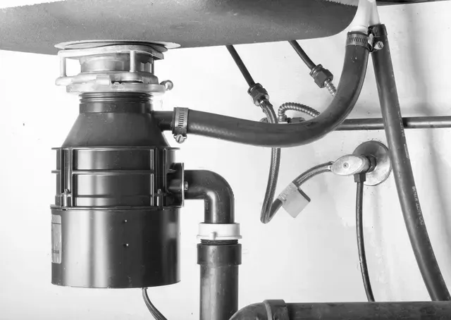 A black garbage disposal unit installed under a kitchen sink, connected to plumbing pipes and a dishwasher hose.