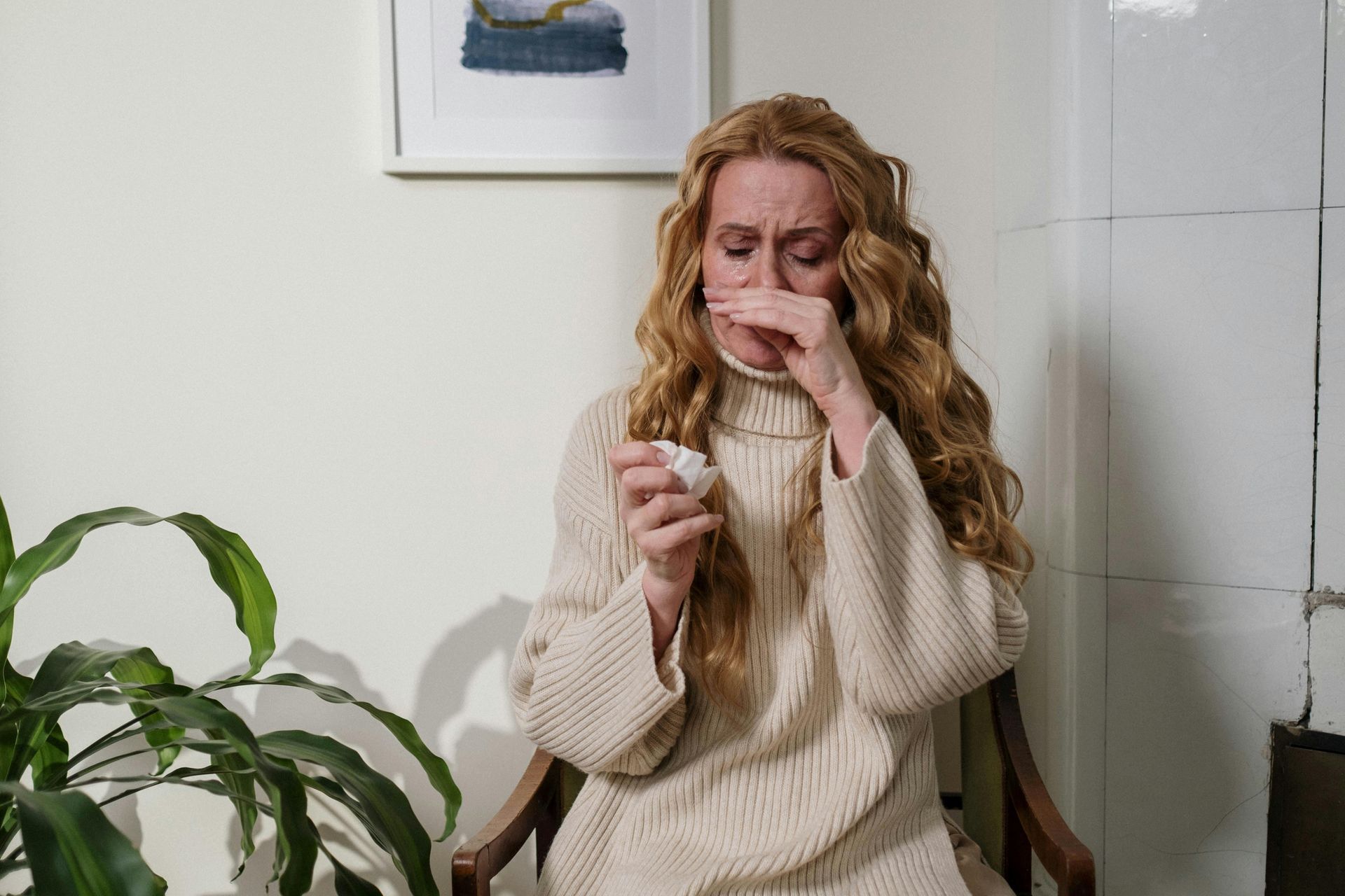A person with long, wavy hair wearing a beige sweater, holding a tissue and covering their nose as if about to sneeze.