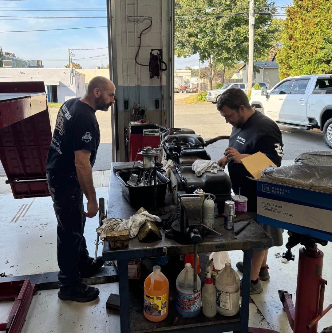 Two mechanics working on an engine in a garage. One examines the engine while the other checks papers.