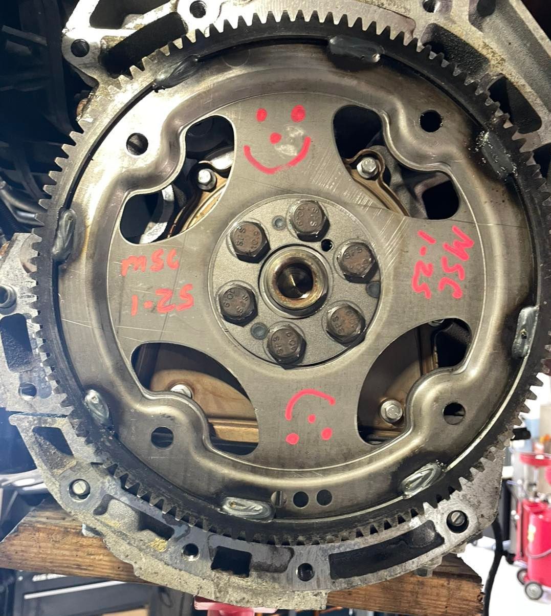 Close-up of a car flywheel with visible teeth. Red smiley faces are drawn on the central metal plate.