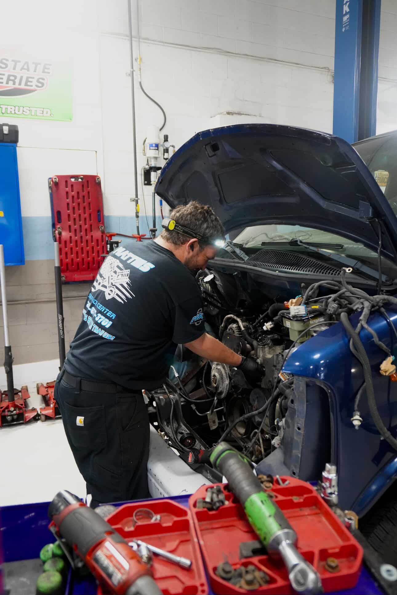 Mechanic working on a blue vehicle with the hood open in a garage, tools visible.