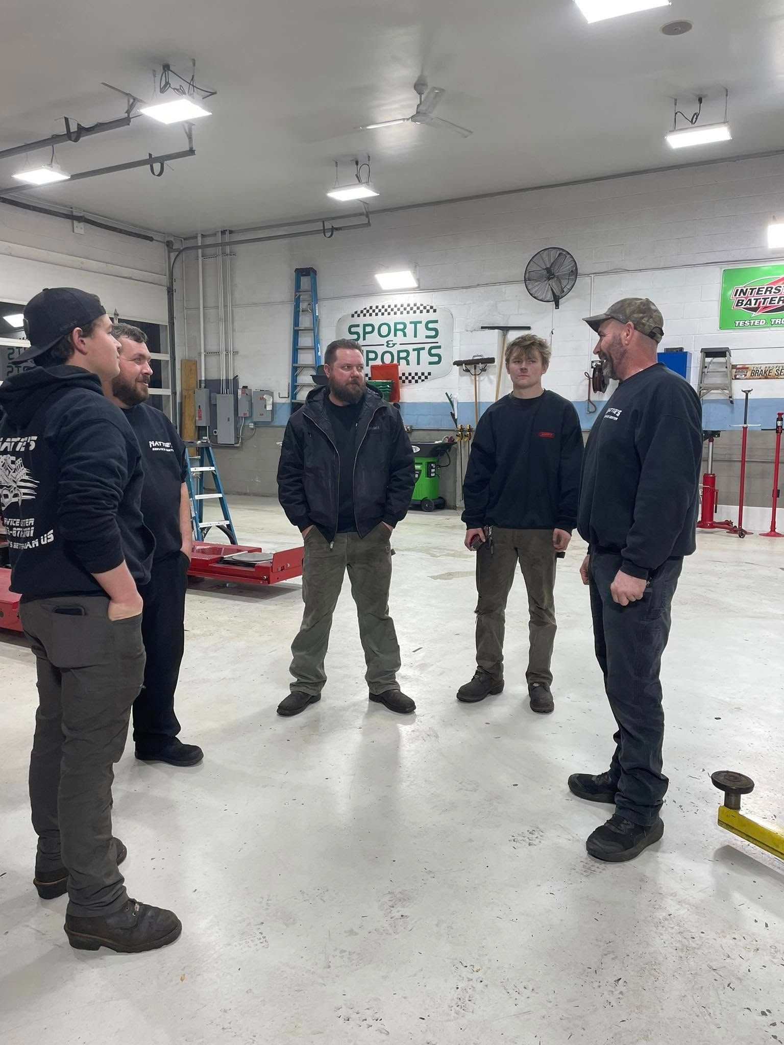 Five men in a shop setting, talking. One man wears a baseball cap, another a beanie.