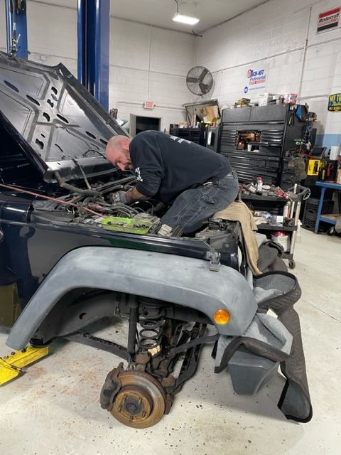 Mechanic working on a Jeep with the hood open in a garage. Front wheel is missing, with rusty brake.