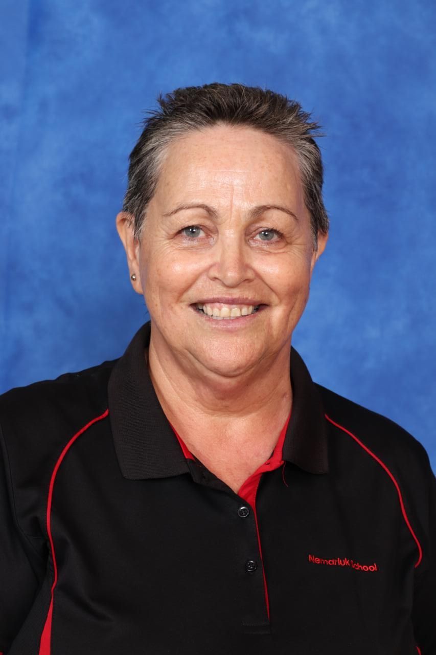 A woman wearing glasses and a red apron is smiling in front of a blue background.