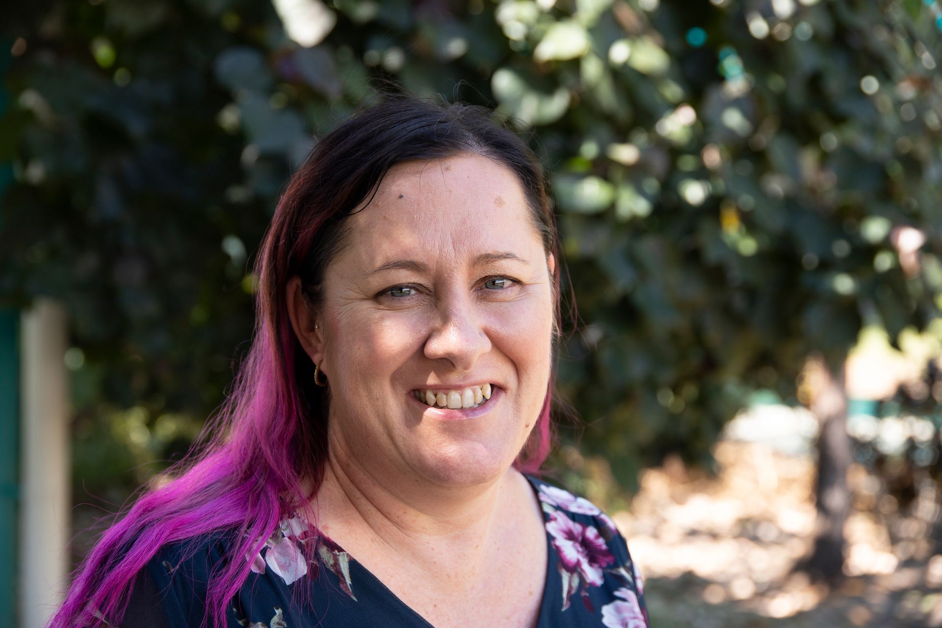 A woman with purple hair is smiling for the camera while standing in front of a tree.
