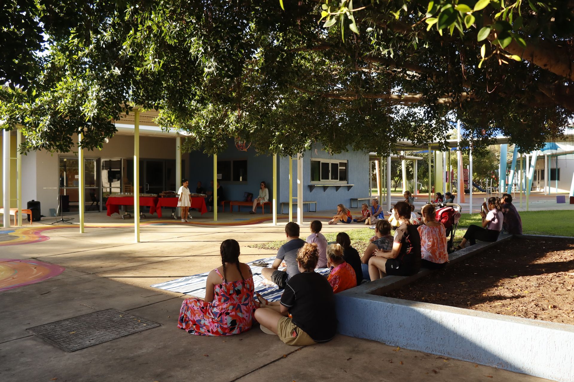 A group of people are sitting under a tree in front of a building.