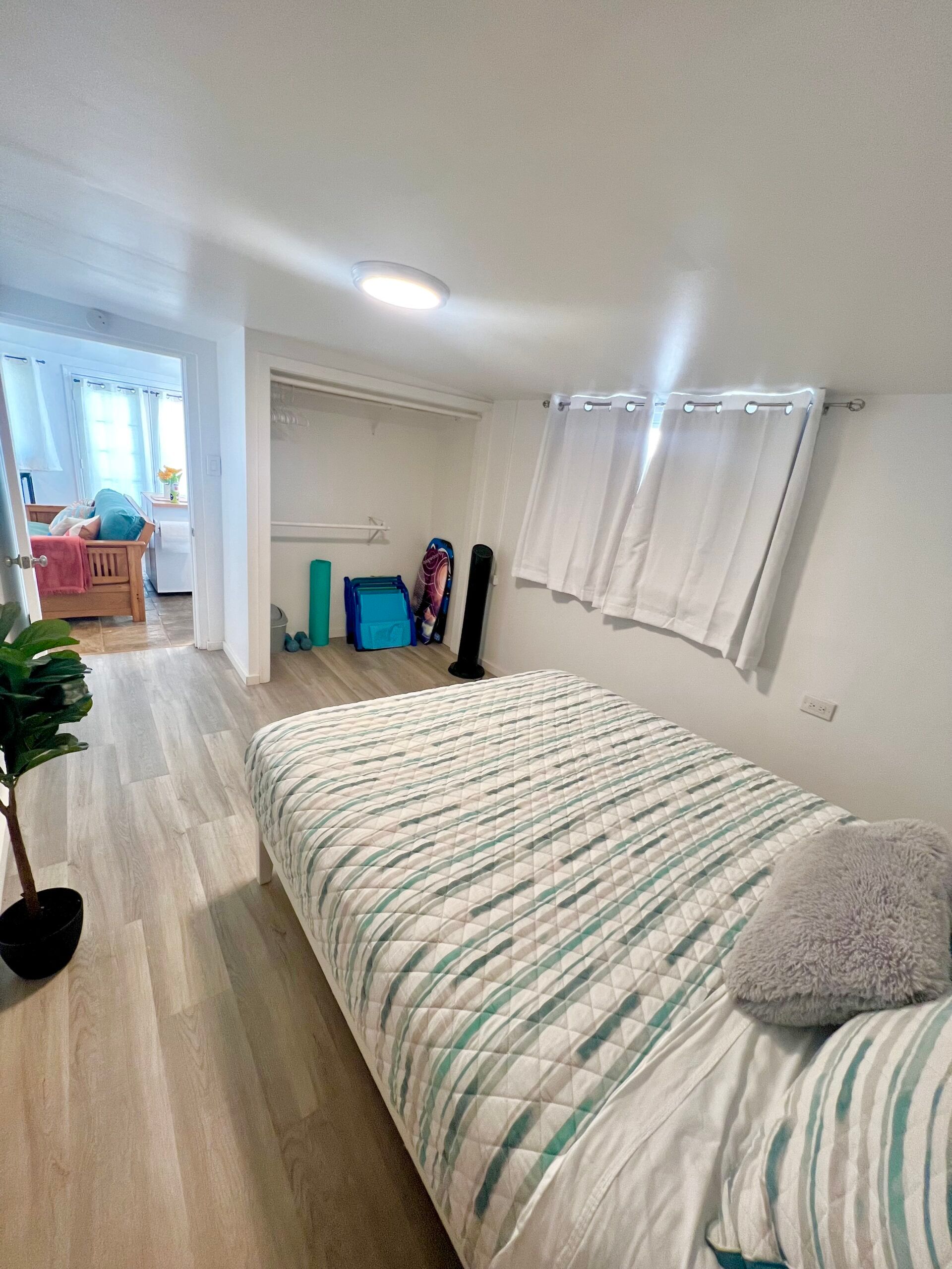 Bedroom with a bed covered in a blue and white striped quilt, adjacent to a hallway with a closet and a small plant.