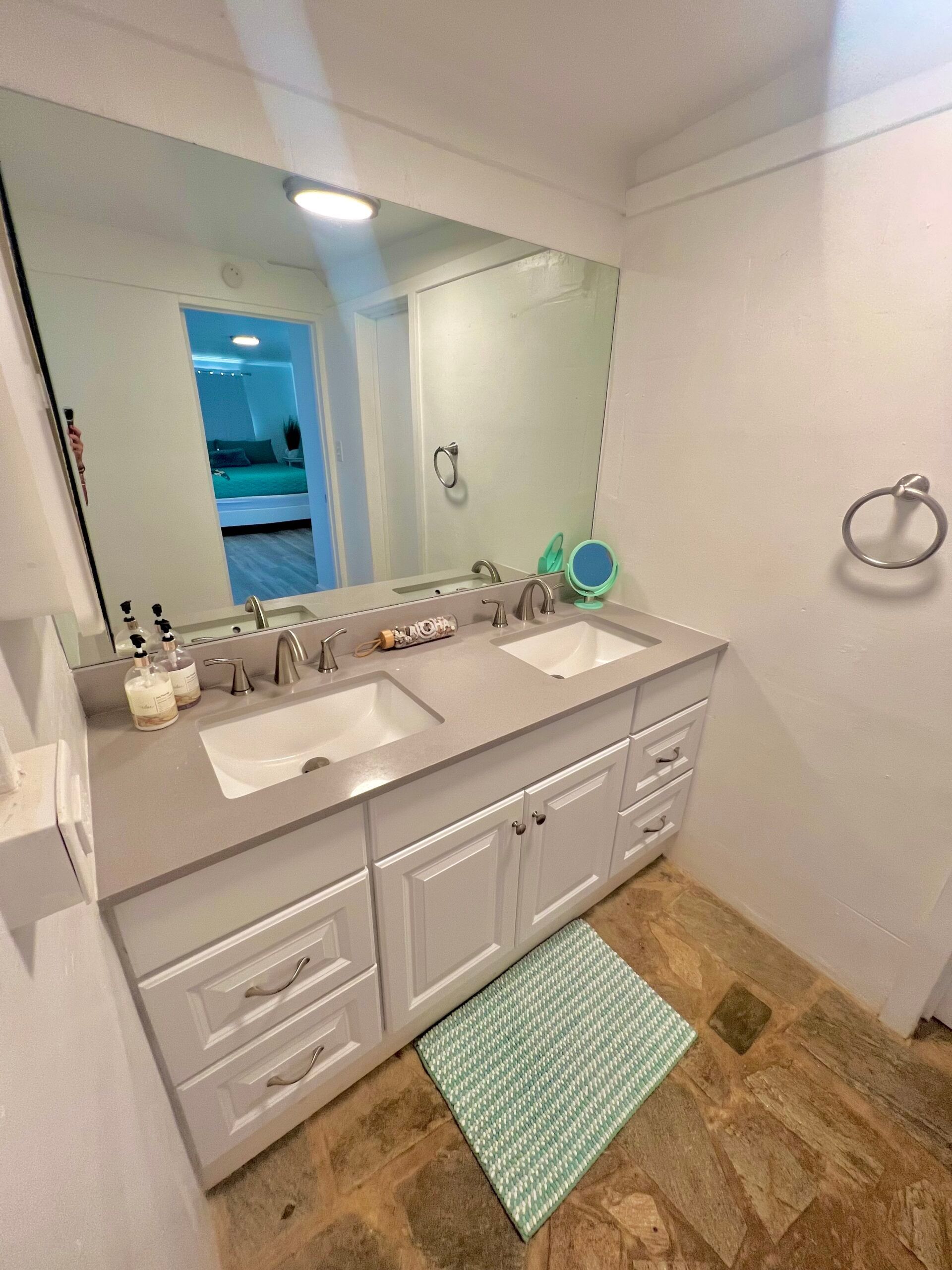 Bathroom with white double vanity, large mirror, and light blue accent rug on brown tile floor.