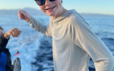 A man wearing sunglasses is standing on a boat holding a fish.