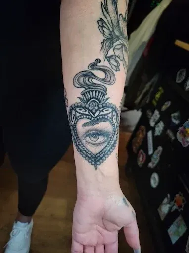 Tattoo of a heart-shaped locket on a forearm, with an eye inside, surrounded by ornamental details and smoke.