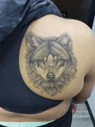 Wolf tattoo on a person's upper back, in shades of gray, with one blue eye.