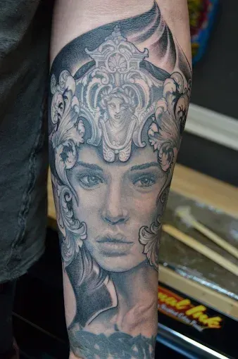 Detailed black and gray tattoo on an arm, portrait of a woman with ornate headdress.