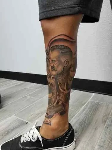 Tattoo of a man's face and abstract designs on a leg, with black and gray ink. The person wears black shoes and gray shorts.