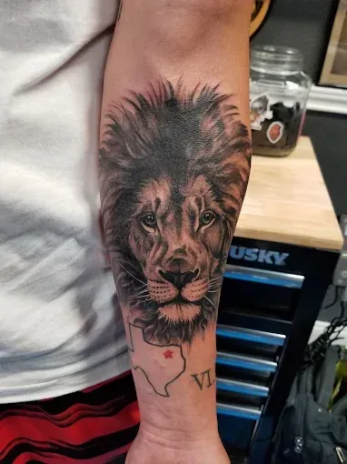 Tattoo of a lion's head on an arm, with a Texas outline and text. Black and gray shading.