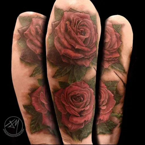 Tattoo of red roses with green leaves on an arm. Dark background.