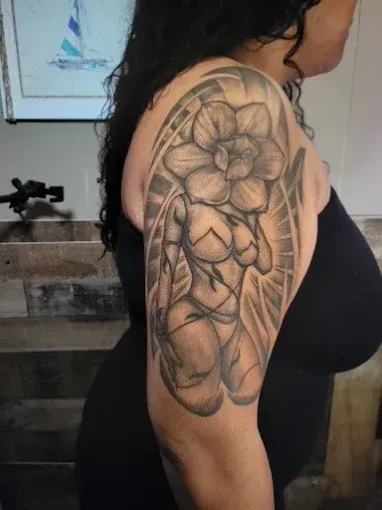 Woman with a detailed black and grey tattoo of a flower-headed figure on her upper arm.