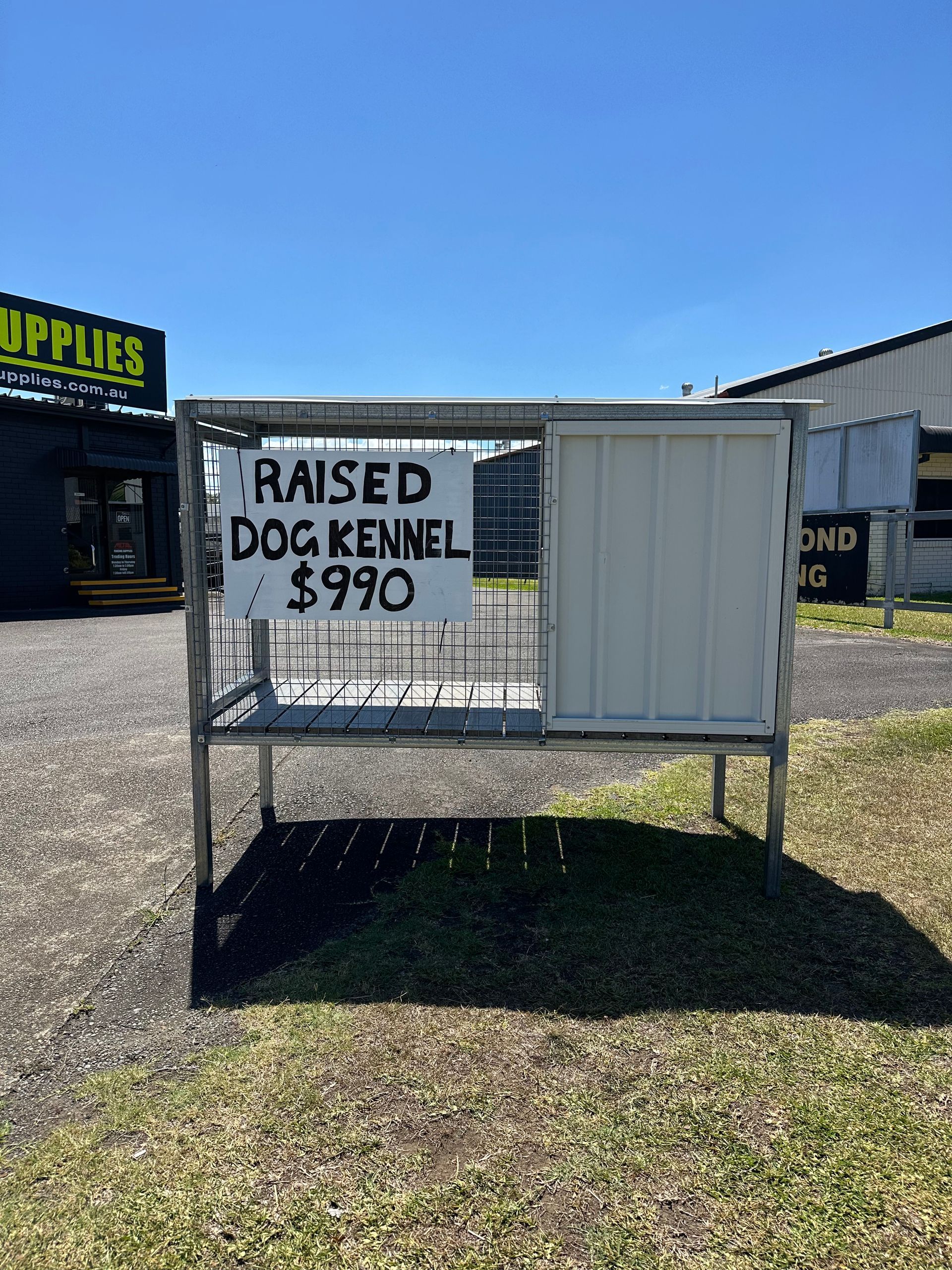 A metal dog kennel for sale, $990. Sign attached. Outdoors on grass, sunny day. Building in background. — Metal Fencing Supplies In Taree, NSW