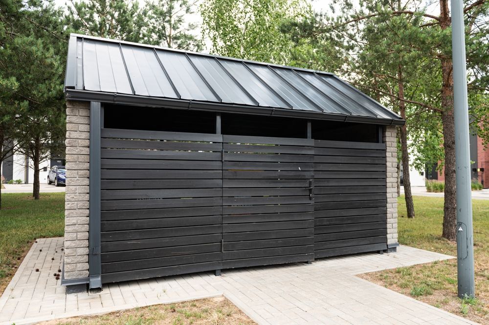 Gray Wooden Trash Enclosure With Black Roof and Brick Sides on a Paved Walkway — Metal Fencing Supplies In Forster, NSW