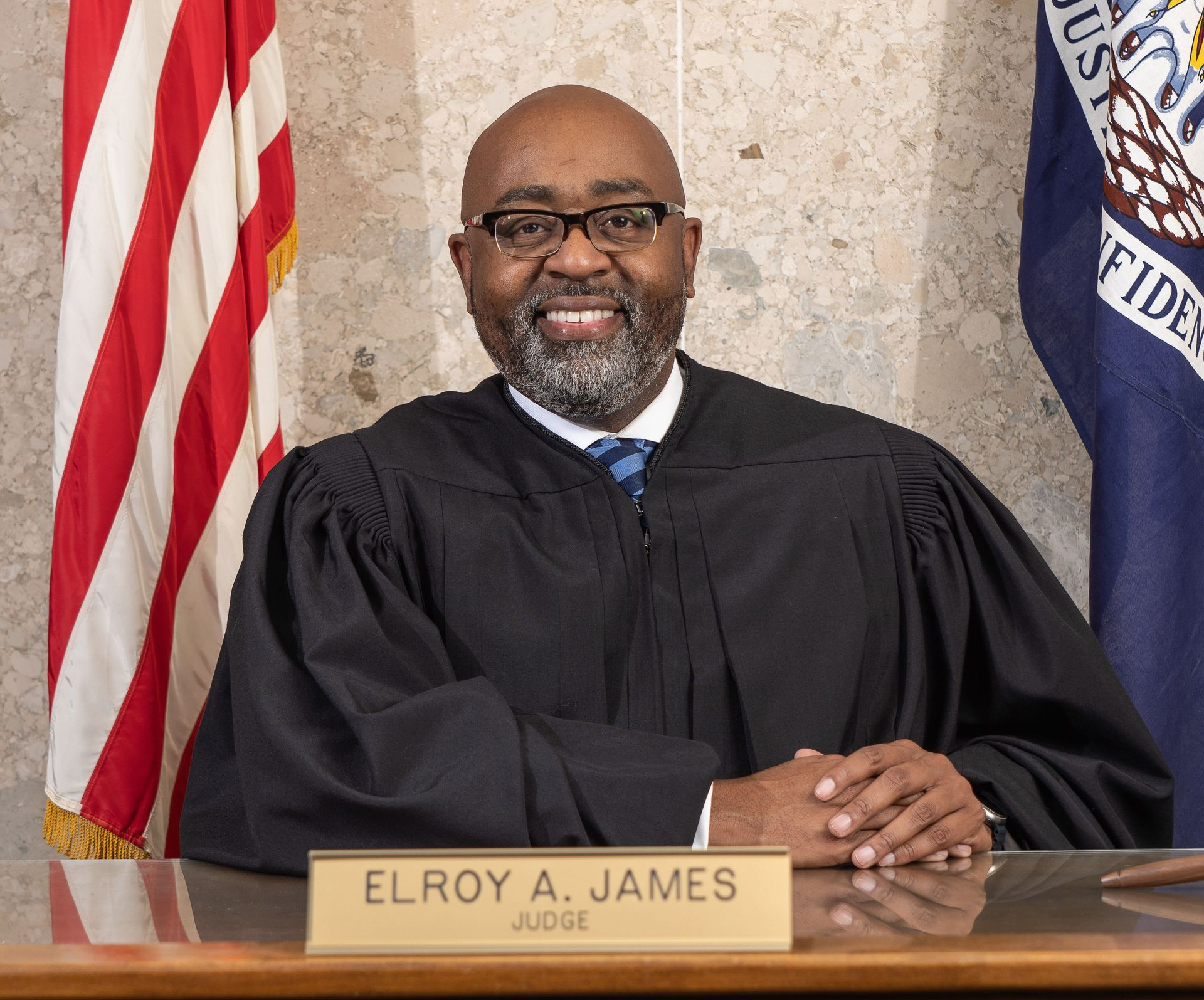 Judge Elroy A. James seated at desk, wearing judicial robes. Smiling in a courtroom setting with flags.