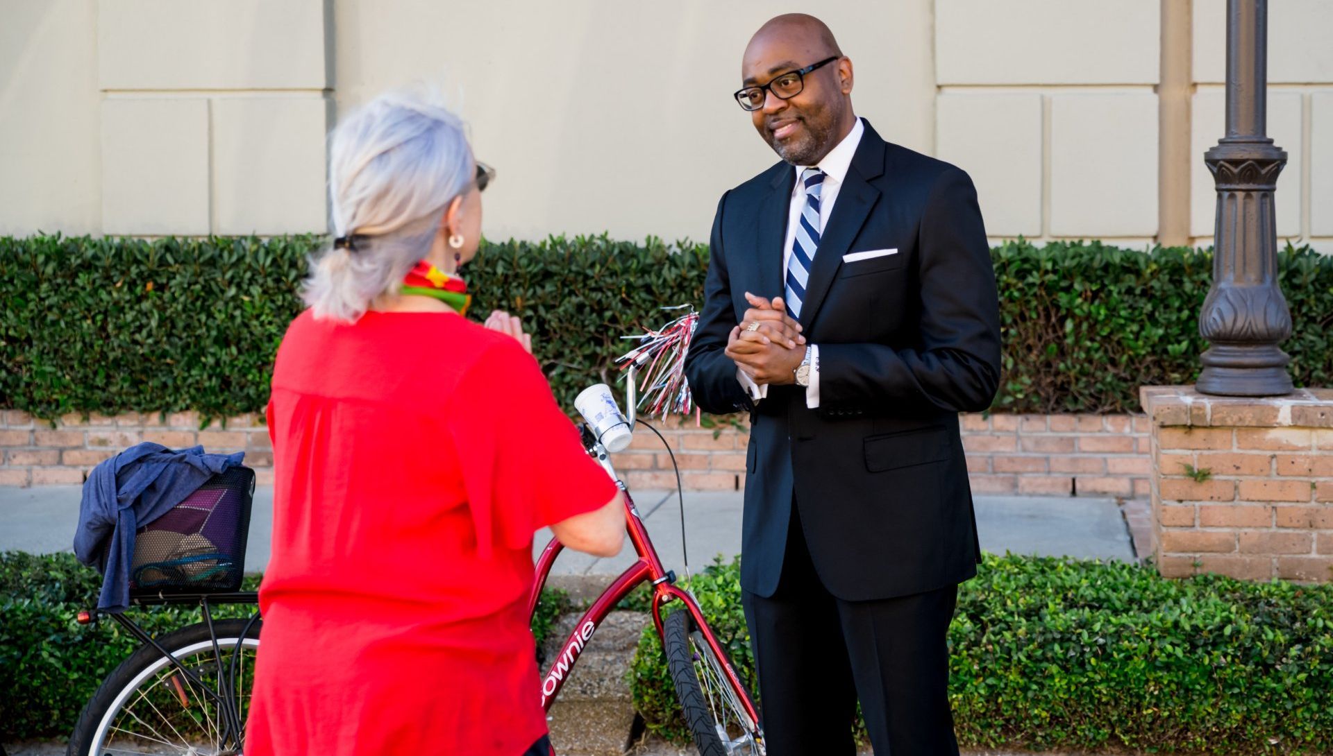 Man in suit talking to a woman in red; bike in between them. Outdoors, daytime.