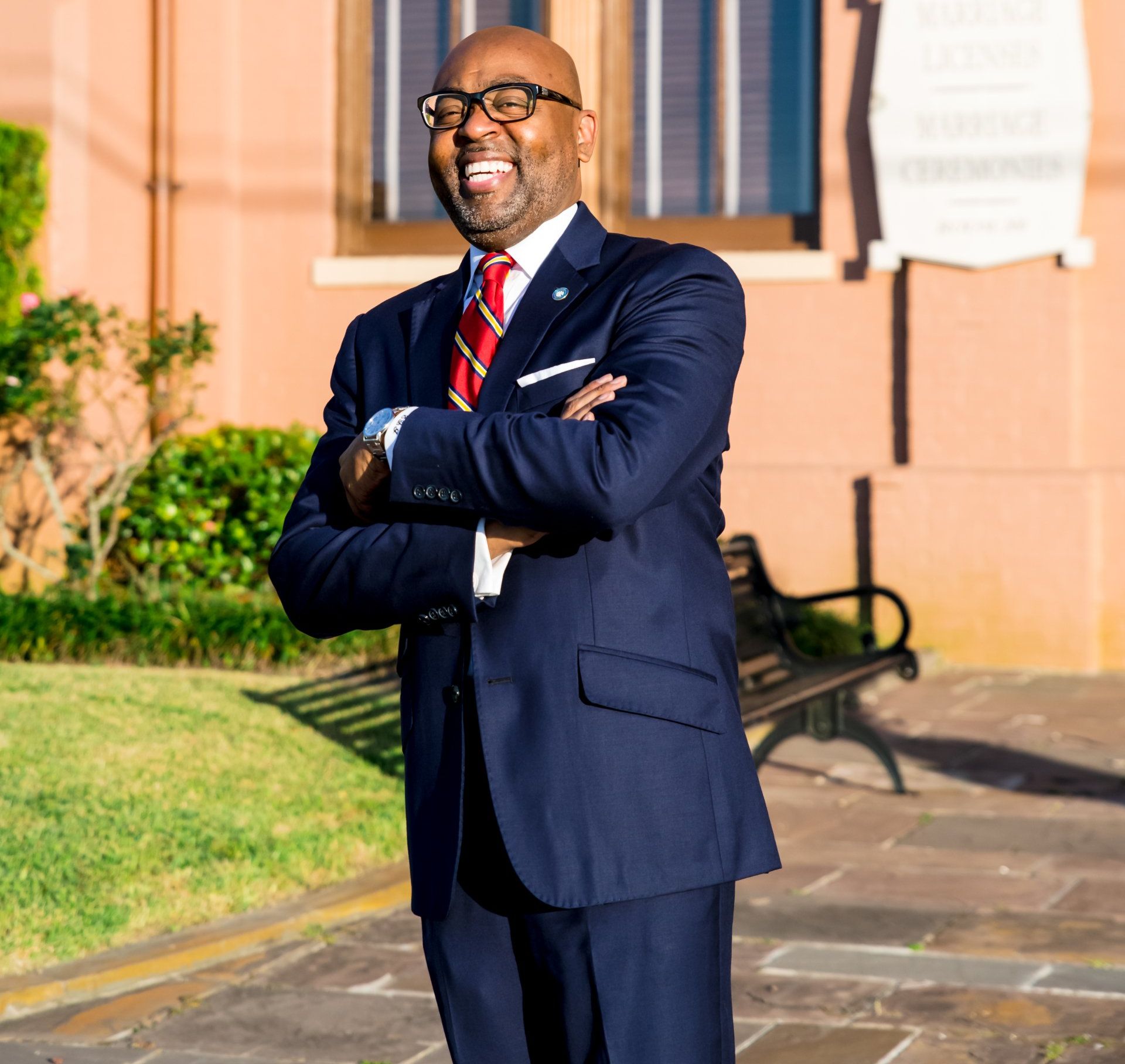 Man in suit, smiling, arms crossed, standing outside a building with a sign.