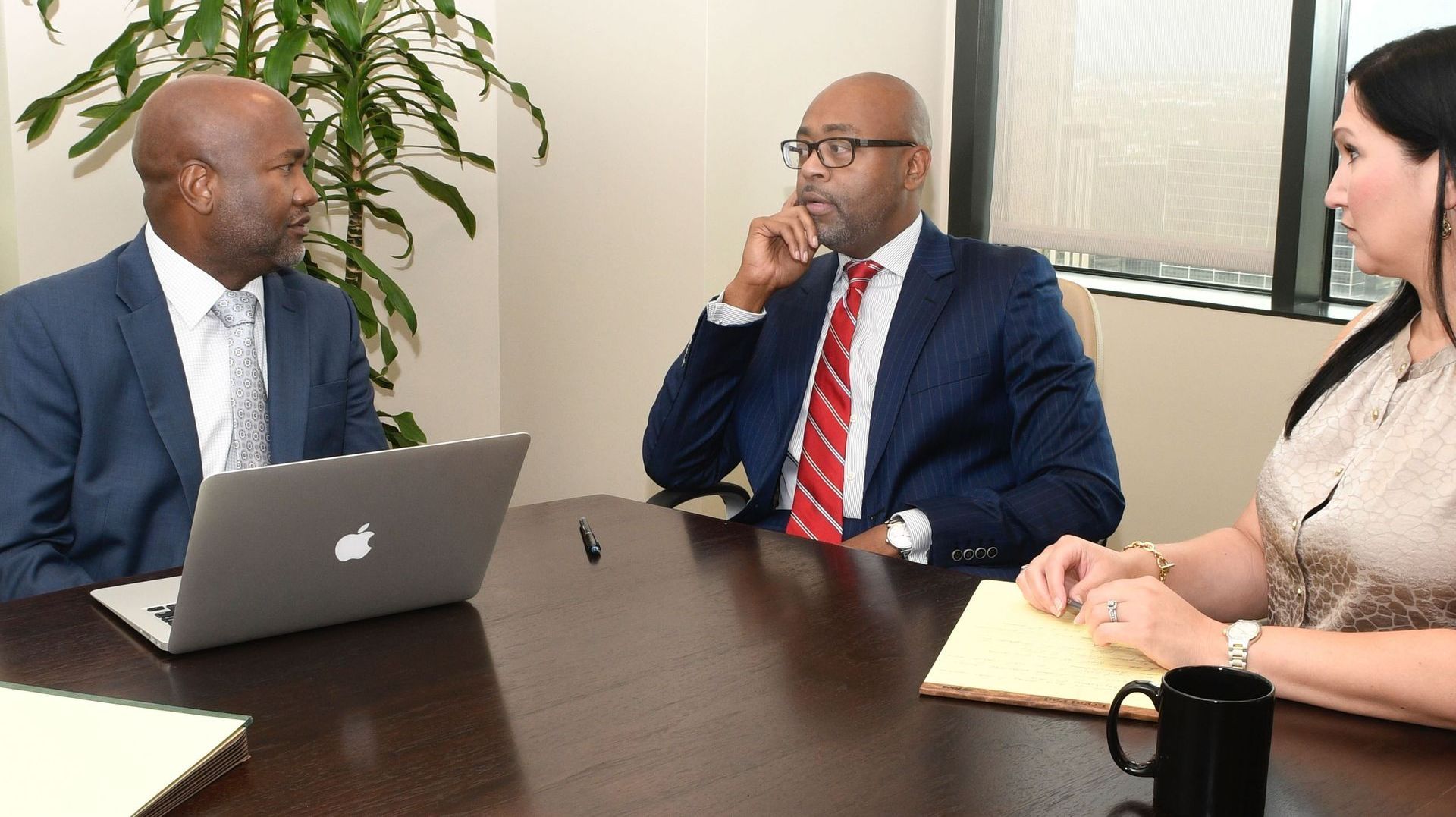 Three people in business attire at a table, discussing with a laptop.