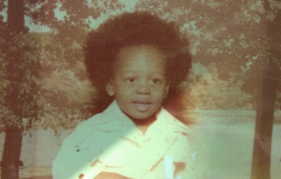 Young Black child with a large afro smiles outdoors wearing a light-colored shirt.