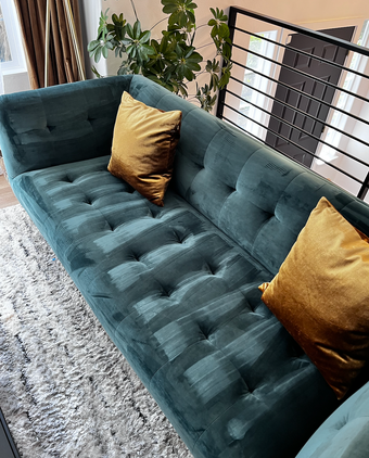 A teal tufted velvet sofa with two gold decorative pillows, positioned near a houseplant and a black metal railing.