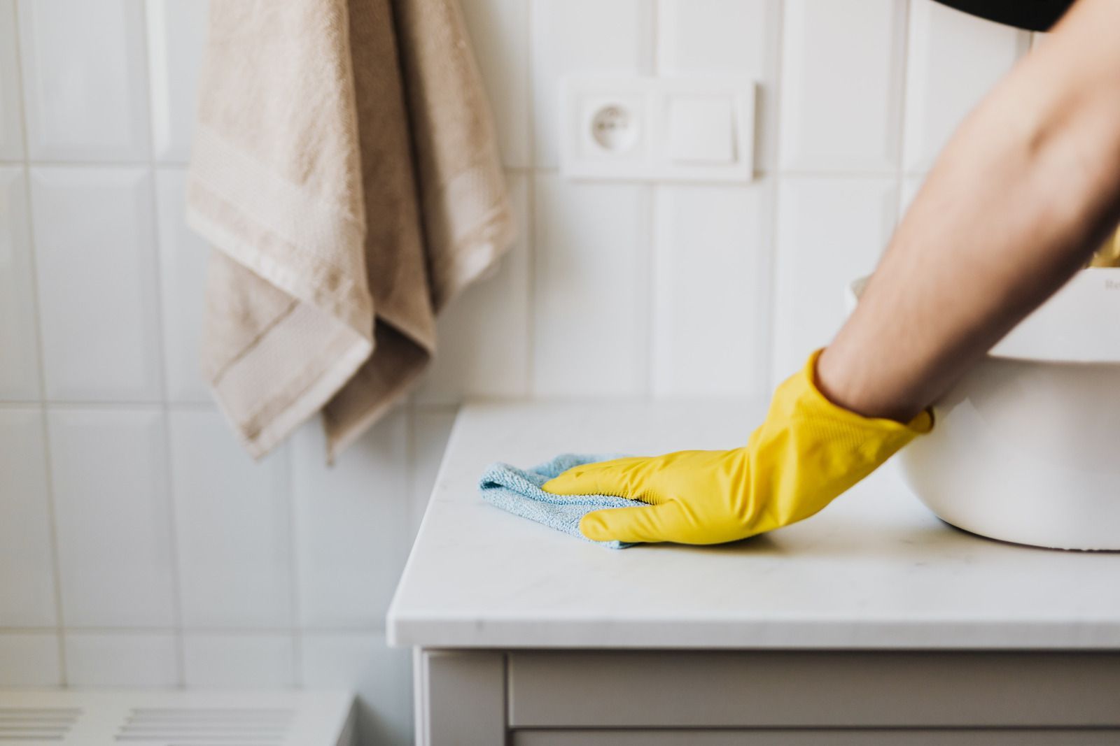 A hand in a yellow rubber glove wipes a white countertop in a tiled bathroom.