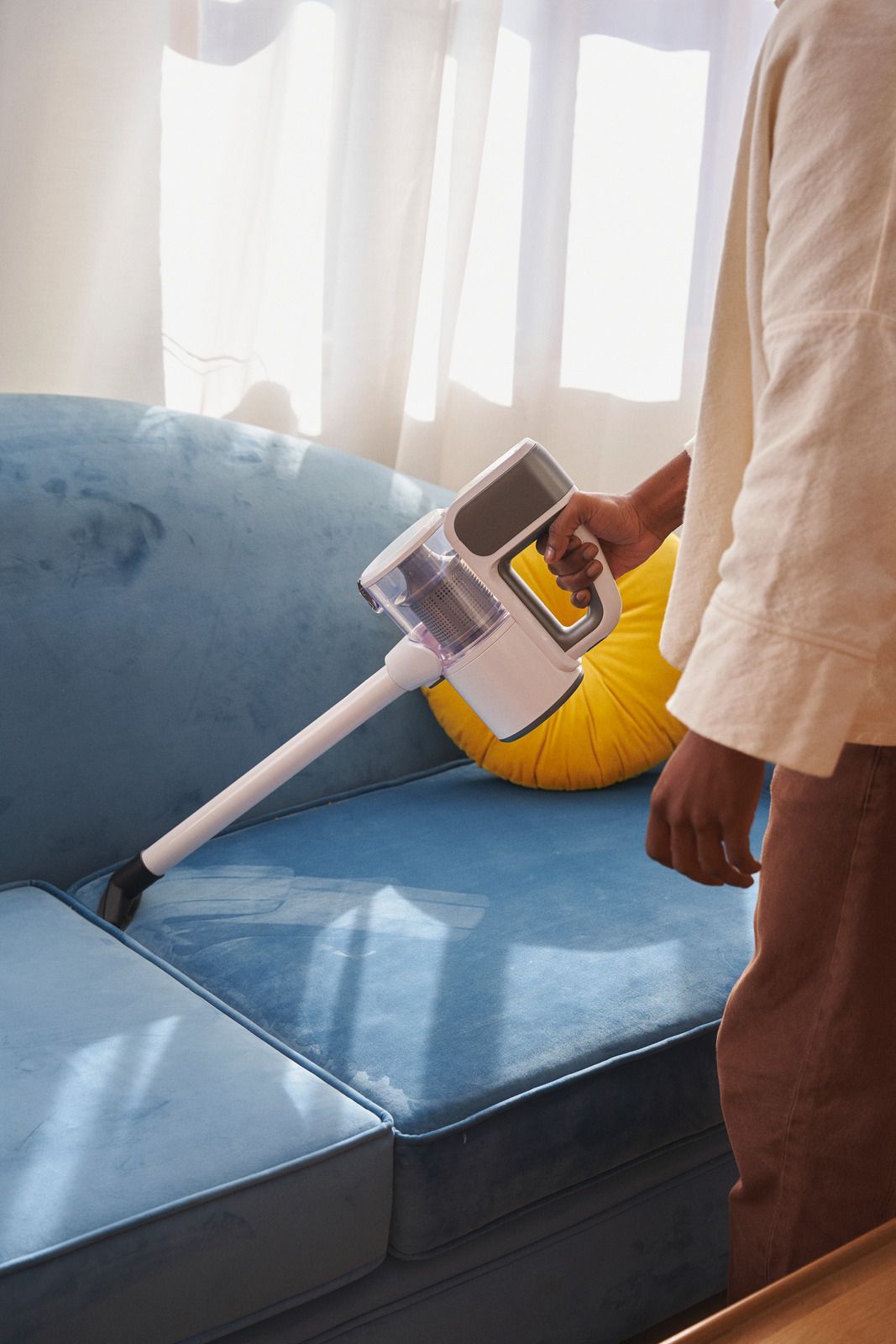 A person vacuums a blue fabric sofa with a white handheld vacuum cleaner near a yellow cushion and bright window.