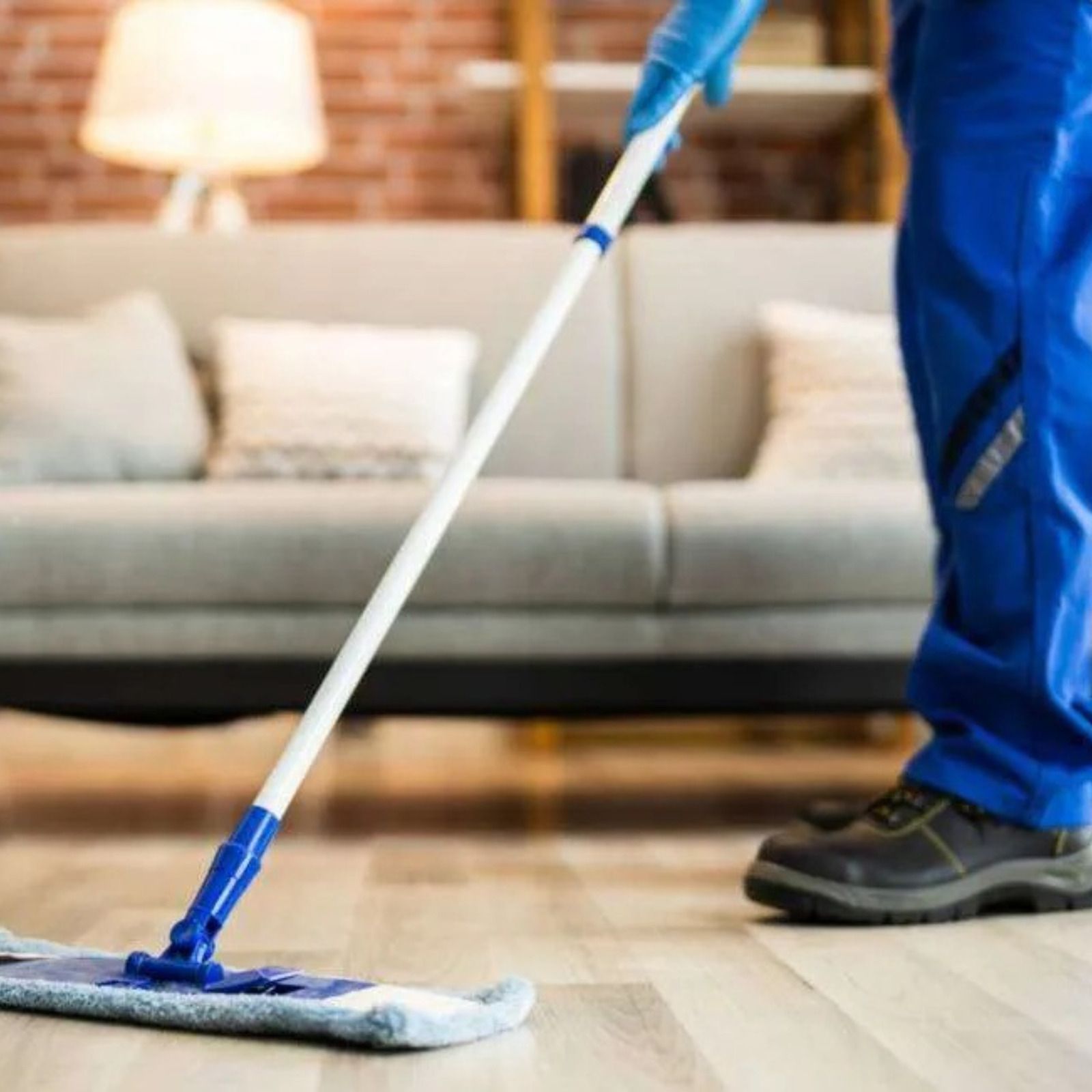A person in blue work clothes and gloves mops a wooden floor in front of a grey sofa.