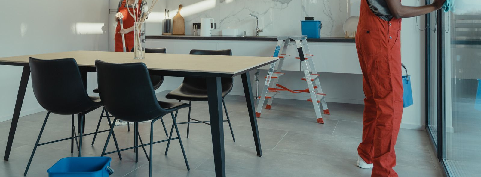 Two people in orange jumpsuits clean a modern kitchen, using a step ladder, bucket, and cloth.