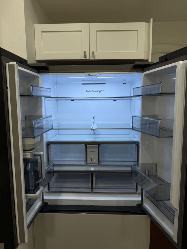 Open empty refrigerator with white interior, glass shelves, and drawers, set below white kitchen cabinets.