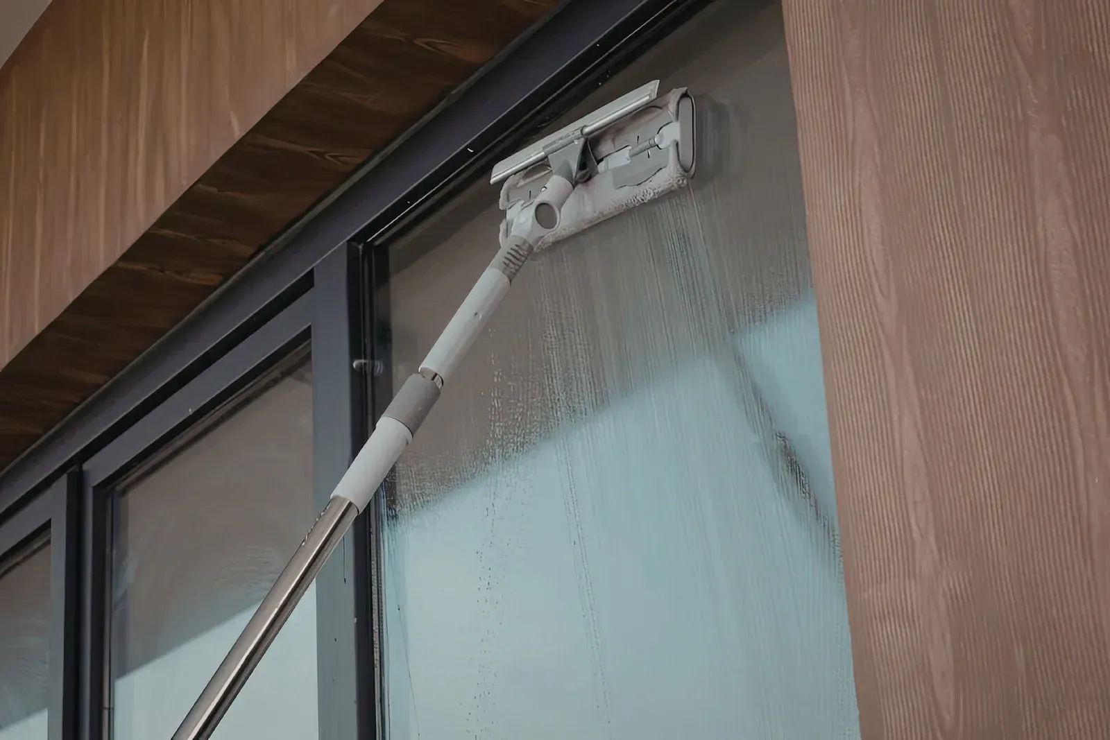 A telescoping pole with a squeegee attachment cleaning a glass window pane with water streaks.