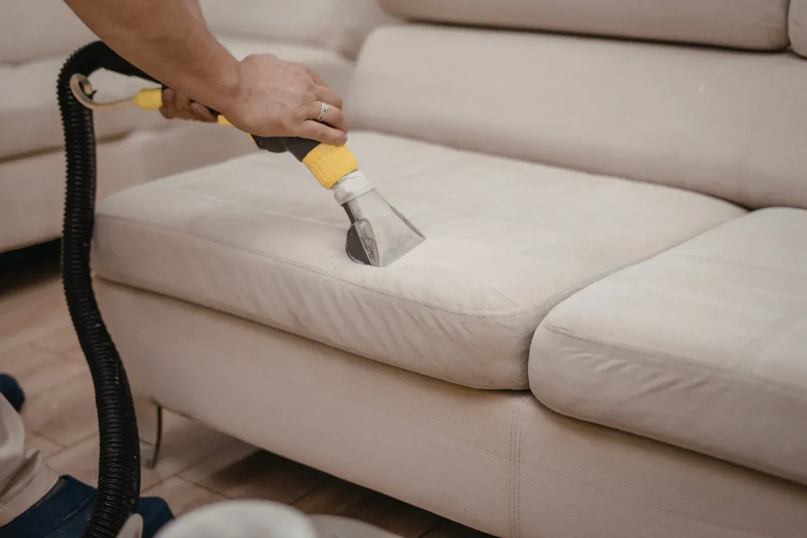 A person uses an upholstery cleaning vacuum tool to deep clean a light-colored fabric sofa.