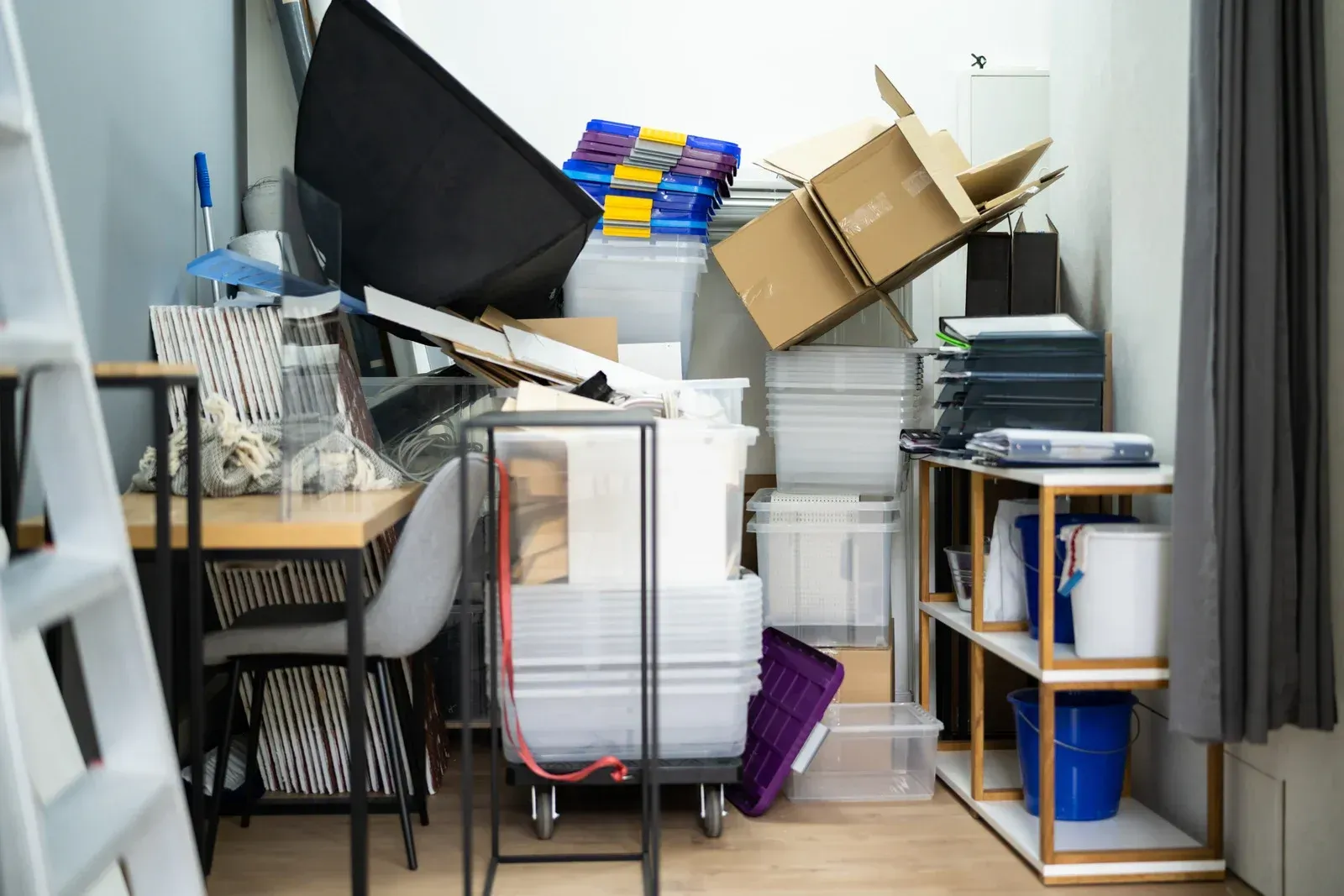 A cluttered storage area filled with stacked cardboard boxes, clear plastic bins, office furniture, and miscellaneous items.