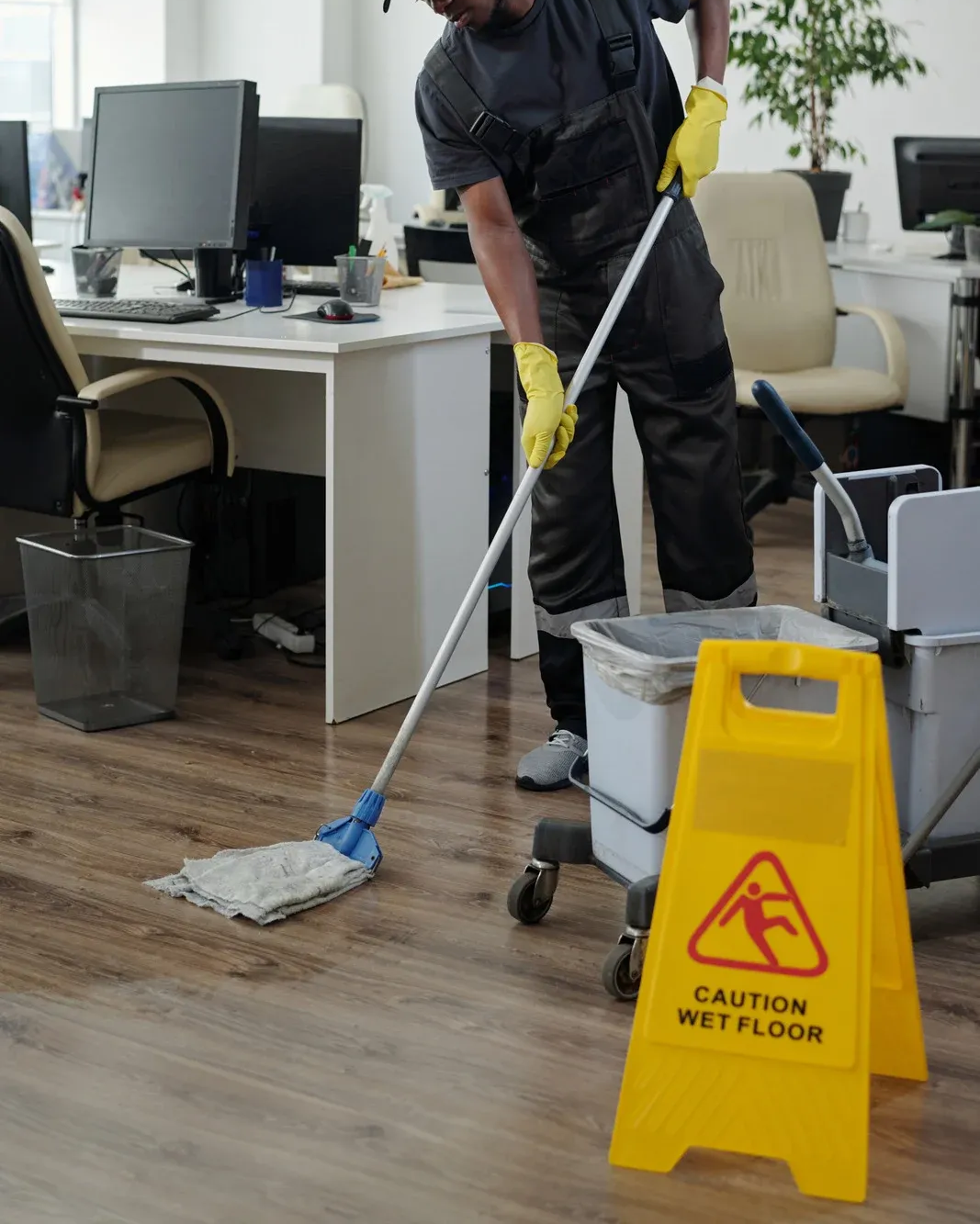 A person in work uniform and yellow gloves mops an office floor near a yellow 
